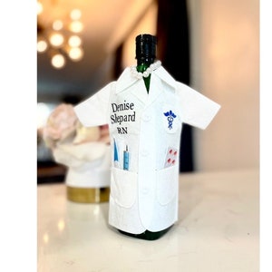 May include: A white lab coat bottle cover with the name Denise Shepard RN printed on the front. The coat has a pocket with a syringe and a blue medical tool. There is a caduceus symbol on the right side of the coat.