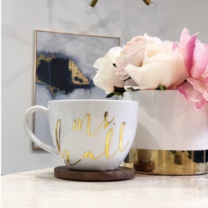 K&ouml;nnte beinhalten: Eine wei&szlig;e Keramiktasse mit einer goldenen Aufschrift "mrs hall". Die Tasse steht auf einem Untersetzer aus Holz. Im Hintergrund steht eine wei&szlig;e Keramikvase mit goldfarbenem Rand, gef&uuml;llt mit rosa und wei&szlig;en Blumen.