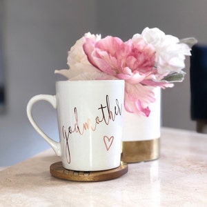May include: White ceramic mug with a gold inscription that reads "godmother" with a small heart below the text. The mug is sitting on a wooden coaster. The mug is next to a white vase with pink flowers.
