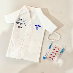 May include: A white doctor's coat with the name "Denise Shepard RN" embroidered on the left side. The coat has a blue medical symbol patch on the right side. A white pearl necklace, a blue thermometer, a pack of red pills, and a syringe are also pictured.