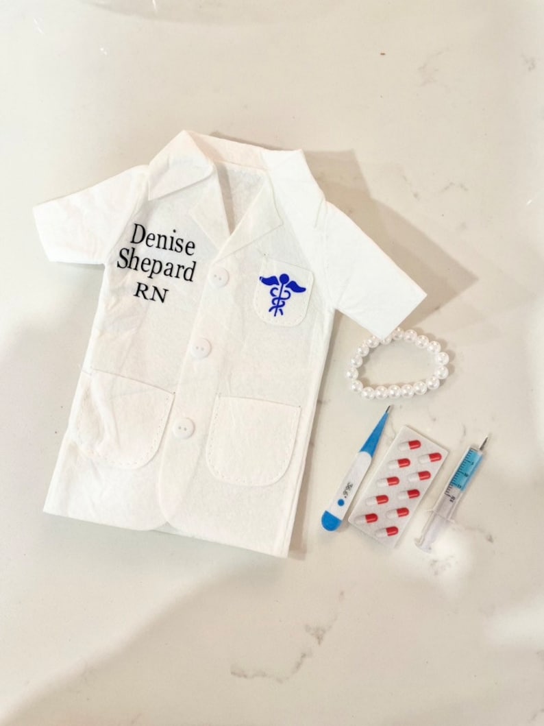 May include: A white doctor's coat with the name "Denise Shepard RN" embroidered on the left side. The coat has a blue medical symbol patch on the right side. A white pearl necklace, a blue thermometer, a pack of red pills, and a syringe are also pictured.