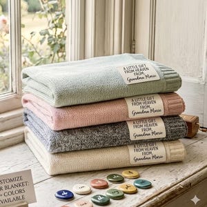 May include: A stack of four folded knit blankets in sage green, soft pink, heather gray, and cream. Each blanket features a white rectangular patch with the black printed text: A LITTLE GIFT FROM HEAVEN FROM, Grandma Marie. Small colored stones are below.