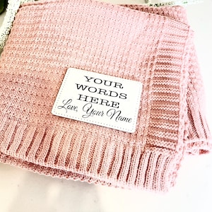 May include: A pink knitted blanket with a white tag that says "YOUR WORDS HERE, Love, Your Name".