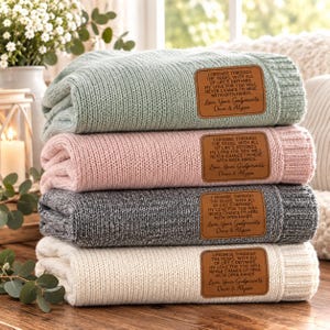 May include: A stack of four folded knit blankets in sage green, soft pink, charcoal gray, and cream. Each blanket features a rectangular brown leather patch with the text: I promise through the years, with all of life's demands, my love for you will never chance, I'm here with open hands. Love, Your Godparents Dave & Alyssa.