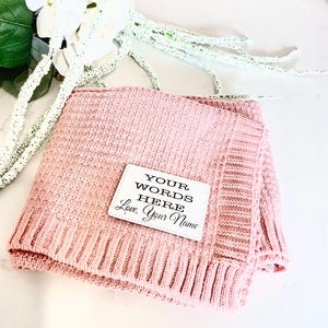 May include: A pink knitted throw blanket with a white square tag that says "YOUR WORDS HERE Love, Your Name"