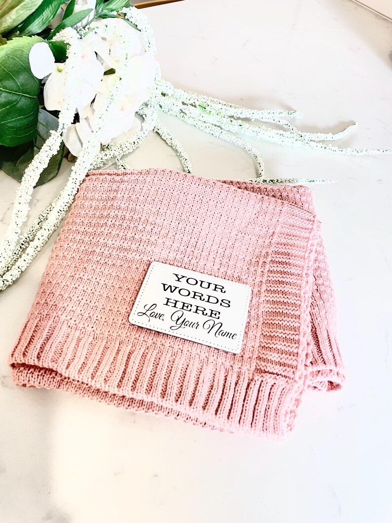May include: A pink knitted throw blanket with a white square tag that says "YOUR WORDS HERE Love, Your Name"