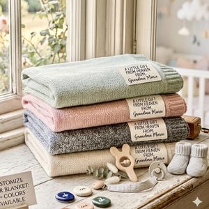 May include: A stack of four folded knit blankets in sage green, soft pink, heather gray, and cream. Each blanket features a rectangular fabric label with the text A LITTLE GIFT FROM HEAVEN FROM, Grandma Marie. Includes a wooden rattle and booties.