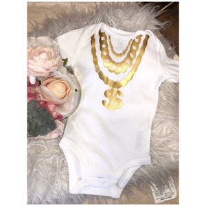 May include: White baby onesie with a gold chain and dollar sign graphic. The onesie is made of a soft, comfortable material and is perfect for a baby shower gift.