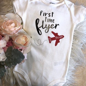 May include: White baby onesie with black text that reads "first time flyer" and a red airplane with the text "Noah 2019" on it.