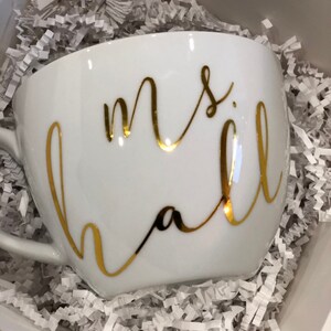 K&ouml;nnte beinhalten: Eine wei&szlig;e Keramiktasse mit goldener Aufschrift "mrs hall". Die Tasse steht in einer Schachtel, die mit wei&szlig;em zerkn&uuml;lltem Papier gef&uuml;llt ist.
