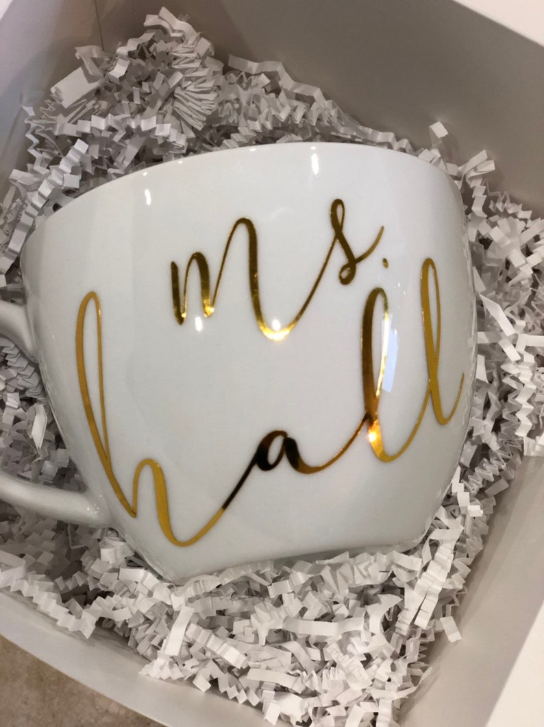 K&ouml;nnte beinhalten: Eine wei&szlig;e Keramiktasse mit goldener Aufschrift "mrs hall". Die Tasse steht in einer Schachtel, die mit wei&szlig;em zerkn&uuml;lltem Papier gef&uuml;llt ist.