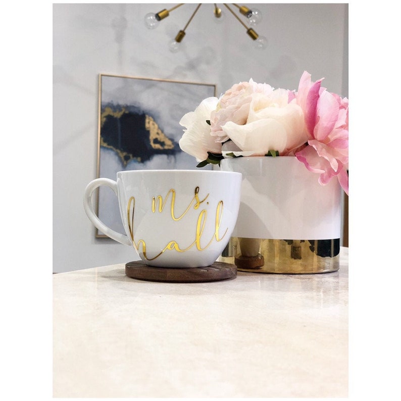 K&ouml;nnte beinhalten: Eine wei&szlig;e Keramiktasse mit einer goldenen Aufschrift "mrs hall". Die Tasse steht auf einem Untersetzer aus Holz. Im Hintergrund steht eine wei&szlig;e Keramikvase mit goldfarbenem Rand, gef&uuml;llt mit rosa und wei&szlig;en Blumen.