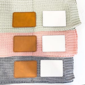 May include: Three knitted blankets stacked on top of each other. Each blanket has a brown leather label and a white label. The blankets are in shades of gray, pink, and light green.