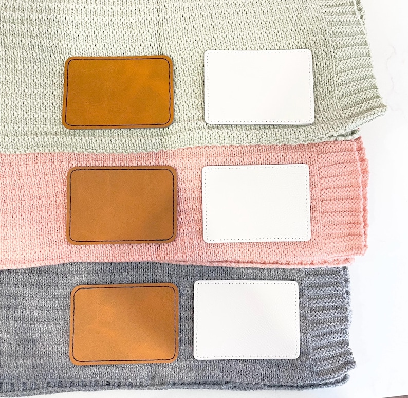May include: Three knitted blankets stacked on top of each other. Each blanket has a brown leather label and a white label. The blankets are in shades of gray, pink, and light green.