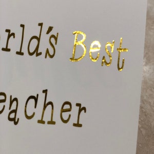 K&ouml;nnte beinhalten: Goldene Folienbeschriftung auf einer wei&szlig;en Karte mit dem Text "World's Best Teacher".