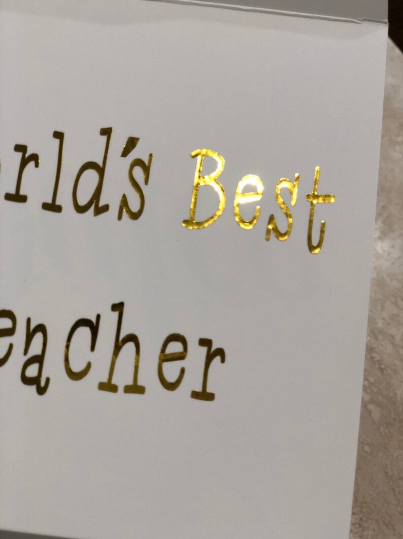 K&ouml;nnte beinhalten: Goldene Folienbeschriftung auf einer wei&szlig;en Karte mit dem Text "World's Best Teacher".