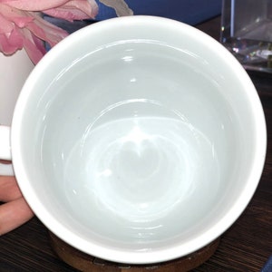 K&ouml;nnte beinhalten: Eine wei&szlig;e Keramiktasse mit einem Untersetzer aus Holz. Die Tasse ist leer und hat eine glatte, gl&auml;nzende Oberfl&auml;che.