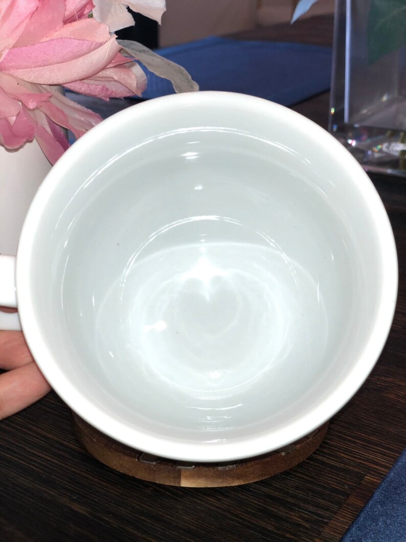 K&ouml;nnte beinhalten: Eine wei&szlig;e Keramiktasse mit einem Untersetzer aus Holz. Die Tasse ist leer und hat eine glatte, gl&auml;nzende Oberfl&auml;che.