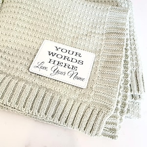 May include: A light green knitted baby blanket with a white tag that says "YOUR WORDS HERE Love, Your Name"
