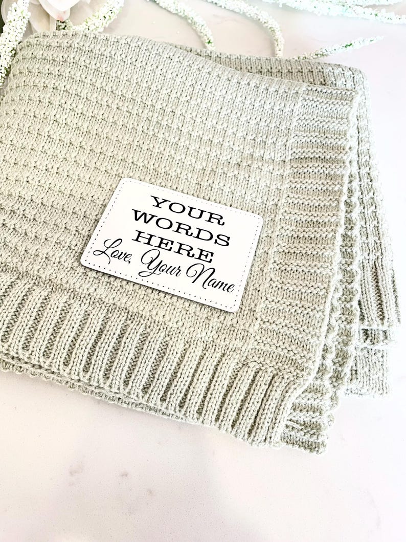 May include: A light green knitted baby blanket with a white tag that says "YOUR WORDS HERE Love, Your Name"