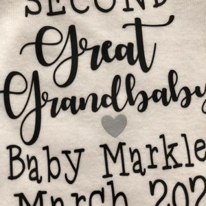 May include: White onesie with black text that reads "Second Great Grandbaby Baby Markle March 2022". A small gray heart is to the right of the text.