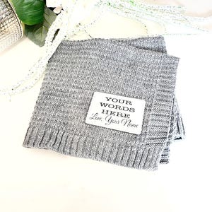 May include: A gray knitted blanket with a white square tag that says "YOUR WORDS HERE Love, Your Name".