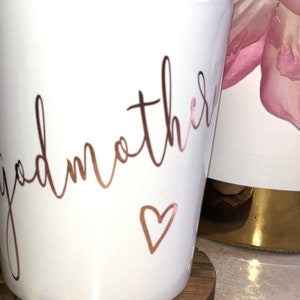 May include: White ceramic mug with the words "Godmother" written in rose gold script and a small rose gold heart.