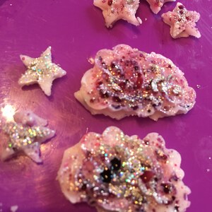 May include: A purple surface with five white star-shaped decorations covered in glitter. Two of the stars are larger than the other three. The larger stars have a pink and white swirl design. The smaller stars are white with a silver glitter design.