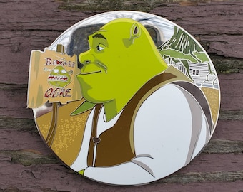 May include: A round, silver-colored pin with a green ogre character from the movie Shrek. The ogre is wearing a white shirt and brown vest. The pin also features a sign that reads "Beware Ogre".