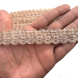 May include: A roll of decorative trim with a wavy, scalloped edge. The trim is a metallic gold colour with a shimmering, textured appearance. The trim is held in a hand against a white background. This trim is suitable for sewing and craft projects.