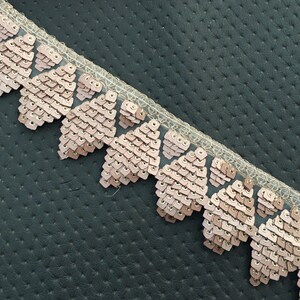 Puede incluir: Primer plano de un ribete de lentejuelas rosa y dorado con borde festoneado. El ribete est&aacute; sobre un fondo texturizado gris oscuro.