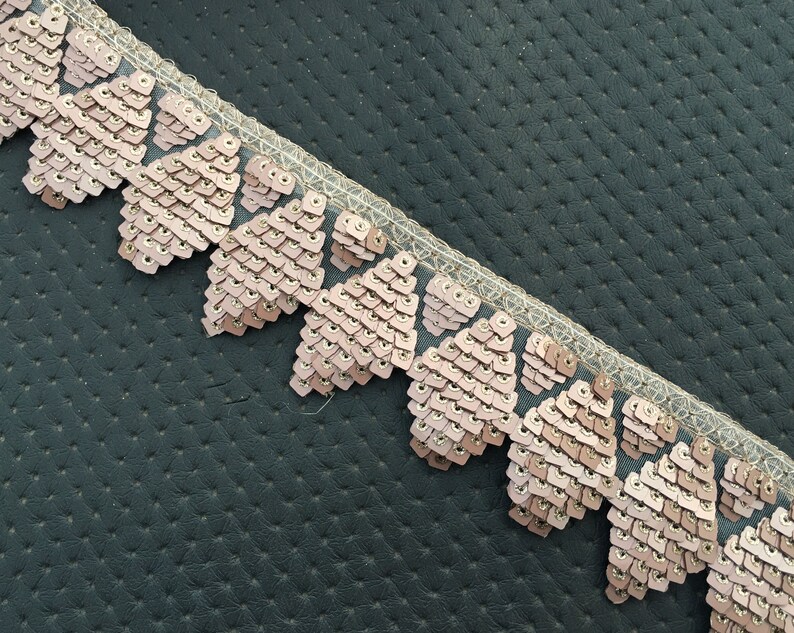 Puede incluir: Primer plano de un ribete de lentejuelas rosa y dorado con borde festoneado. El ribete est&aacute; sobre un fondo texturizado gris oscuro.