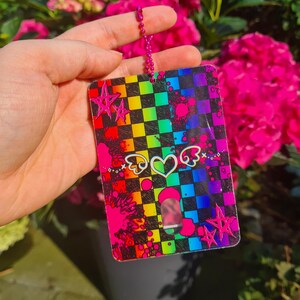 May include: A rectangular, rainbow-checkered tag with a pink chain. The tag features pink star and heart graphics, and pink paint splatters. The background includes vibrant pink flowers.