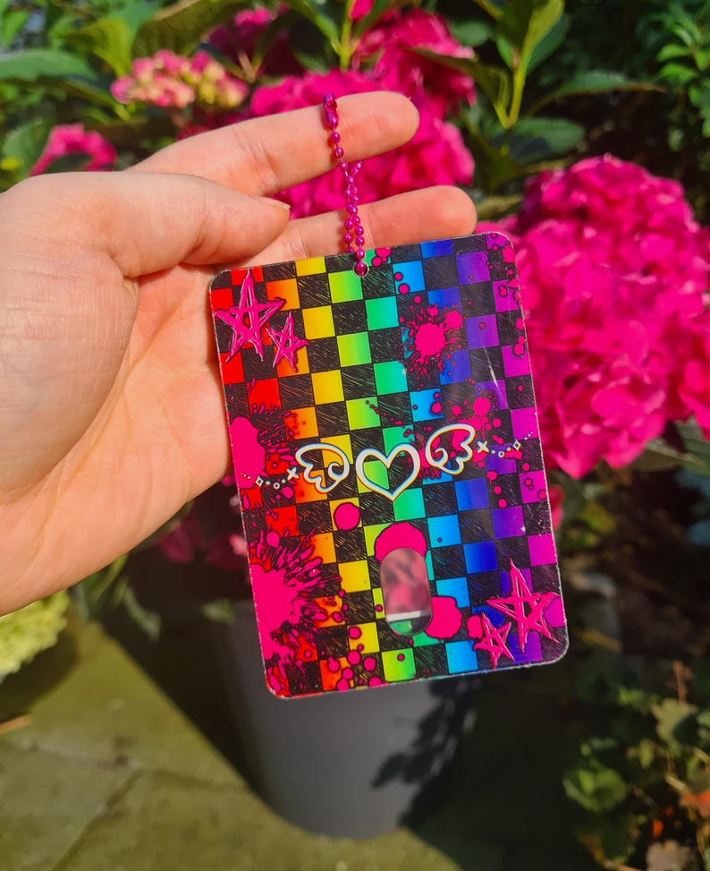May include: A rectangular, rainbow-checkered tag with a pink chain. The tag features pink star and heart graphics, and pink paint splatters. The background includes vibrant pink flowers.