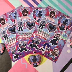 May include: Sticker sheets and heart-shaped badges with cartoon characters and a black and white creature. The stickers have a purple and red marbled background. Text includes "And Demon Loves to All".