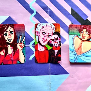 Op de afbeelding: Drie geïllustreerde stickers met cartoonfiguren. De eerste sticker toont een vrouw met lang bruin haar die een rood shirt en een ketting draagt. De tweede sticker toont een man met wit haar die een rood shirt en een pop met een rode strik draagt. De derde sticker toont een man met bruin haar die een blauw shirt draagt.