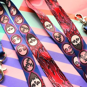 May include: Several lanyards with a repeating pattern of cartoon faces and skulls. The lanyards are primarily blue with red and pink accents. Each lanyard has a metal clasp and a blue fabric neck strap.