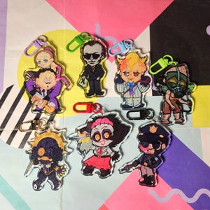 May include: A collection of seven acrylic character keychains featuring diverse illustrated figures, each attached to a colourful plastic lobster clasp. The designs include various stylised individuals in distinct outfits, poses, and vibrant colour palettes.