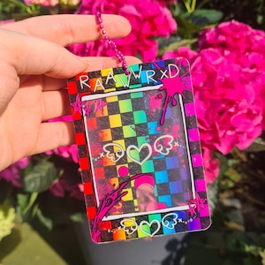 Puede incluir: Un porta-credenciales colorido con un fondo a cuadros de arcoíris y el texto "RAAWRXD". El diseño incluye corazones alados y detalles de salpicaduras rosas. Una cadena rosa está adjunta en la parte superior.