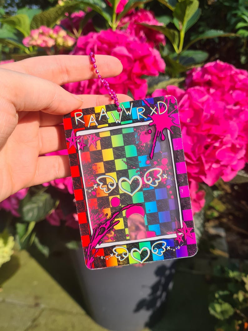 May include: A colorful ID badge holder with a rainbow checkered background and the text "RAAWRXD". The design includes hearts with wings and pink splatter accents. A pink chain is attached to the top.