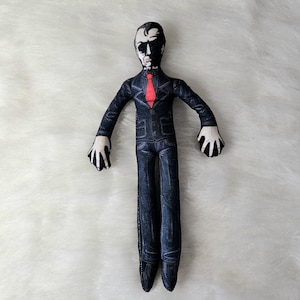 Peut inclure: Une poupée en tissu avec un motif imprimé représentant une personne en costume sombre avec une cravate rouge. La figurine a un visage bicolore avec un côté ombré en noir et l'autre clair, portant une veste et un pantalon noirs avec des coutures visibles.