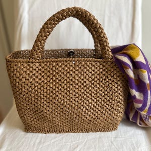 May include: A woven brown tote bag with a braided handle. The bag has a rectangular shape and a button closure. A patterned scarf in purple, yellow, and white is partially visible inside the bag.