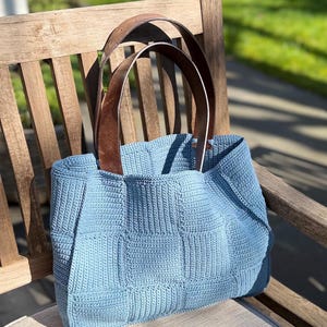 Könnte beinhalten: Hellblaue gehäkelte Tragetasche mit braunen Ledergriffen. Die Tasche hat ein strukturiertes, quadratisches Muster. Die Griffe sind mit braunen Lederriemen an der Tasche befestigt. Die Tasche steht auf einer Holzbank.