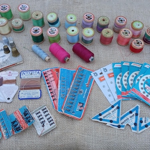 May include: A collection of vintage sewing supplies including various colourful thread spools, Singer hand sewing needles, metal thimbles, assorted buttons on cards, snap fasteners, and a roll of blue ribbon, all arranged together for sewing projects.