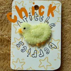 May include: A yellow felt chick with orange beak and feet on a white card with black and orange text that says "little chick" and a pattern of stars.