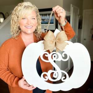 May include: A white pumpkin-shaped door hanger with a burlap bow and a monogrammed letter "S" in a decorative script font.