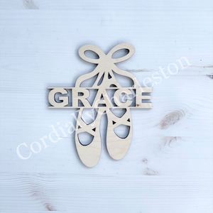May include: Wooden cutout of a pair of ballet shoes with a bow on top. The word "GRACE" is cut out of the shoes.