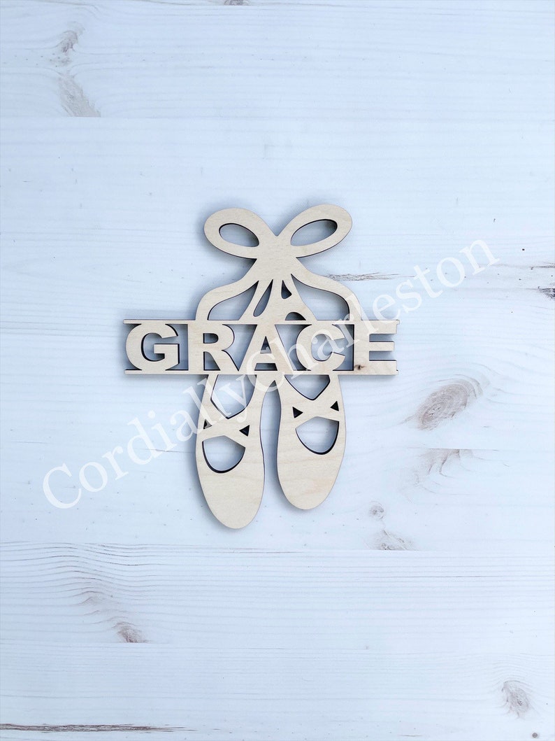 May include: Wooden cutout of a pair of ballet shoes with a bow on top. The word "GRACE" is cut out of the shoes.