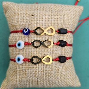 May include: Three red string bracelets featuring a gold-tone infinity symbol, a round evil eye bead, and a small black bead. The top bracelet has a dark blue evil eye bead, while the middle and bottom bracelets have white evil eye beads.