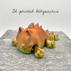 May include: 3D printed kittysaurus, a cute orange and green dinosaur with cat features. The figurine is lying on a wooden surface with the text "the pink penguin" written below it.